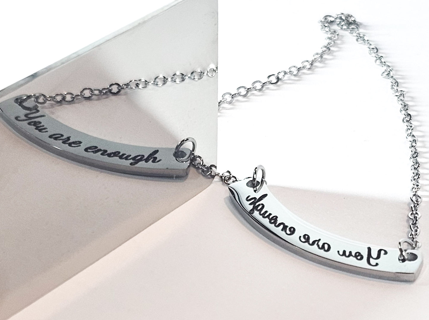 A silver-toned crescent-shaped necklace with the engraved script phrase 'You are enough' on a white background, shown in a split layout with a mirror reflection on the left and the actual product on the right.