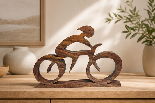 A dark brown wooden sculpture of a cyclist in motion, displayed on a light wood console table in a styled interior setting with a plant and vases in the background.