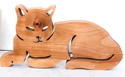 A wooden cat figurine carved from light brown wood, shown lying down on a white surface against a white background.