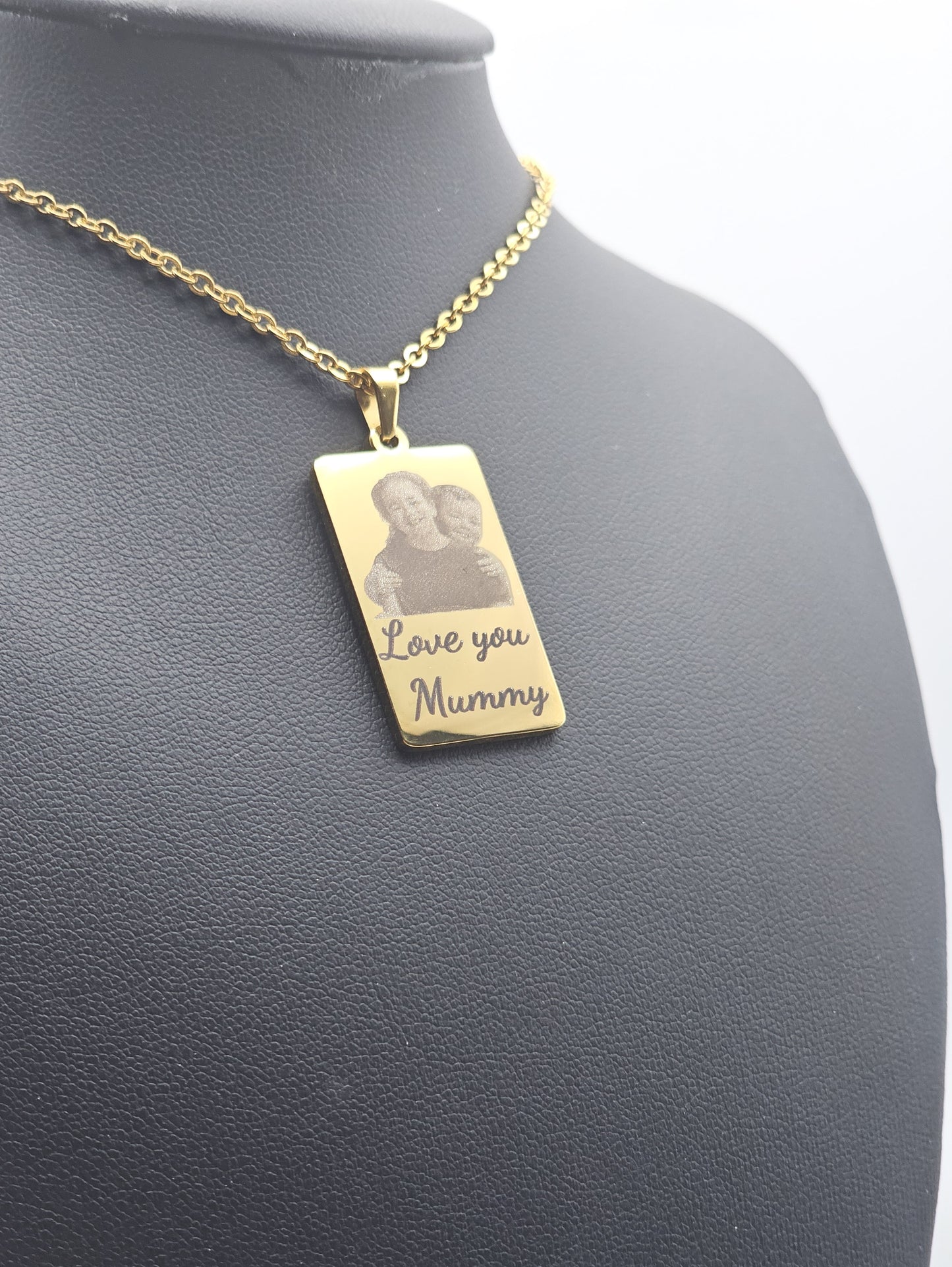 A gold rectangular pendant necklace displayed on a dark gray mannequin bust. The pendant features a custom engraved photo of a two children engraved at the top, with the words 'Love you Mummy' written below in cursive.